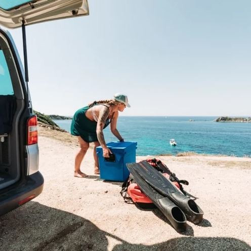 Eine Frau an der Küste hebt eine blaue Kühlbox von Qool auf