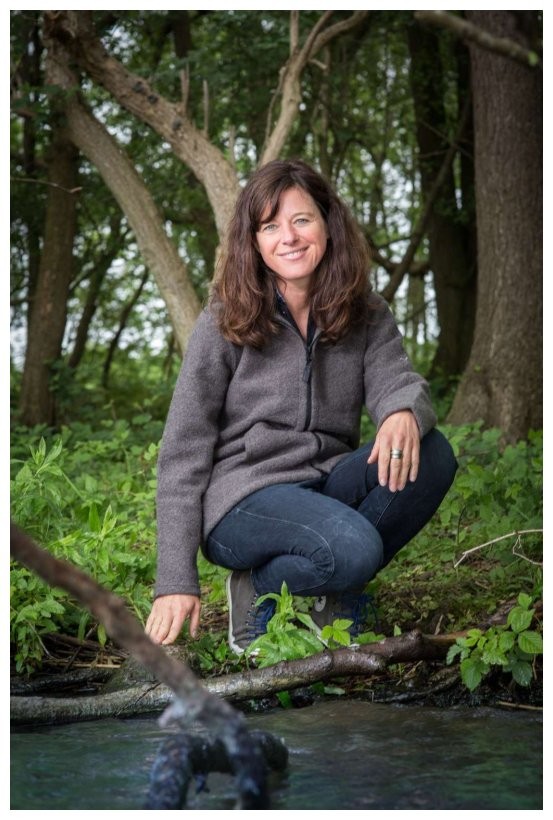 Eine Frau in Mufflon Jacke kniet vor einem Fluss