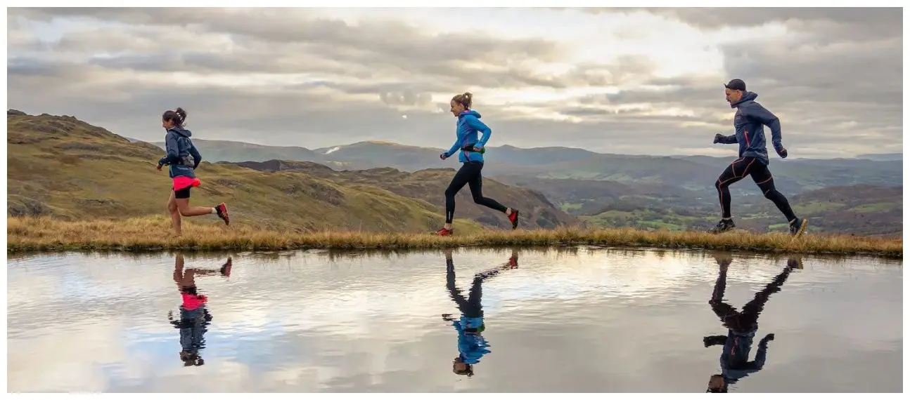 Zwei Frauen und ein Mann joggen entlang eines kleinen Bergsees in OMM Kamleika Bekleidung